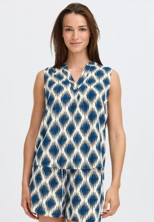 Žena oblečená v bezrukávovej námorníckej modrej a bielo vzorovanej blúzke s krátkymi nohavicami na mieru, stojaca na jednoliatom svetlom pozadí.