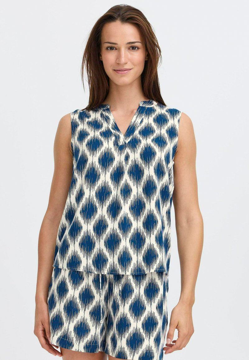 Žena v námořnicky modrém tílku s bílým vzorem a odpovídajících krátkých kalhotách stojí před světlým jednobarevným pozadím.