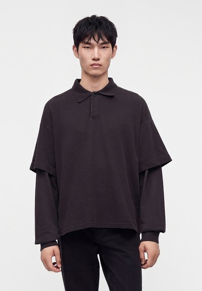 Jeune homme aux cheveux noirs courts portant un haut noir à manches longues ample et superposé ainsi qu'un pantalon noir, debout devant un fond blanc uni.