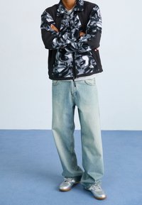Chaqueta de polar con estampado negro y gris sobre una camiseta gris, combinada con jeans holgados de color azul claro y zapatillas plateadas.
