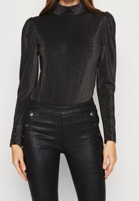 Svart långärmad topp med hög krage och skimrande textur. Figursydd design med puffade axeldetaljer. Matchas med glänsande svarta byxor med knapppdekorationer.