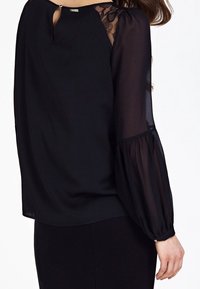 Blusa negra con mangas largas transparentes, detalles de encaje en los hombros y escote en forma de lágrima. Tejido suave con un corte relajado.