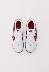 Bele športne copate v beli barvi z rdečimi in črnimi detajli, ki imajo perforirano zasnovo, vezanje na vezalke in gumijaste podplate. Vidna Nike blagovna znamka.