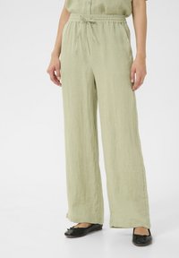 Pantalons en lin vert clair, amples, avec une taille élastique et un cordon de serrage. Associés à des ballerines noires avec un accent de nœud.