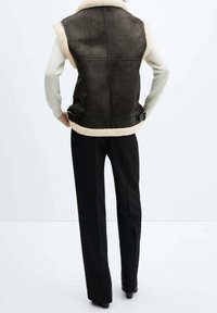 Gilet en simili cuir marron foncé avec col en fausse fourrure crème. Comprend des bretelles latérales réglables et une finition texturée. Pantalon noir.