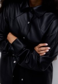 Veste-chemise en similicuir noir avec un devant boutonné, un col pointu et des détails de couture visibles ; elle présente une coupe décontractée et des manches longues.