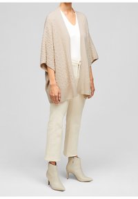 Cardigan beige con texture, dalla vestibilità ampia, con davanti aperto e orlo a coste, indossato sopra una maglietta bianca e abbinato a pantaloni color crema e stivali alla caviglia.
