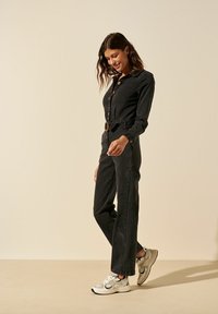 Combinaison en denim noir avec un col, fermeture à boutons sur le devant et taille cintrée avec une ceinture. Associée à des baskets blanches et grises.