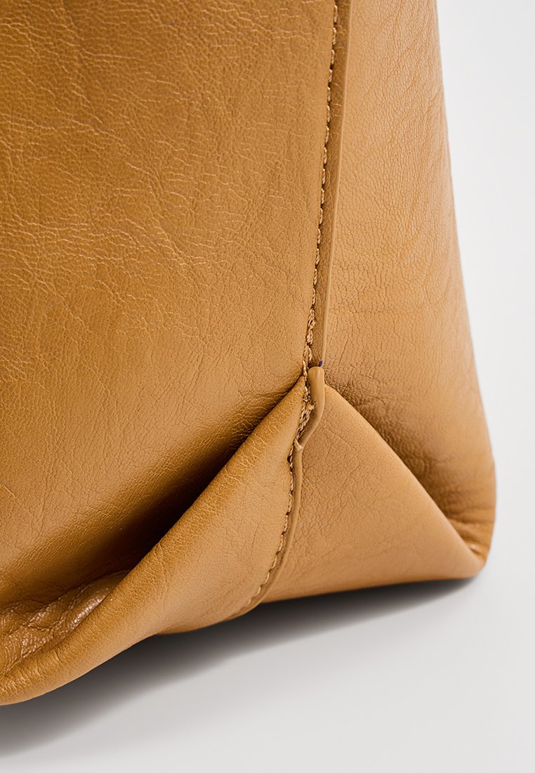 Gros plan sur le coin inférieur d'un sac en cuir beige montrant la surface texturée et la couture détaillée.