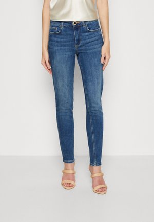 Femme portant un jean skinny bleu, des talons beiges à brides et un haut blanc sans manches, debout devant un fond uni.