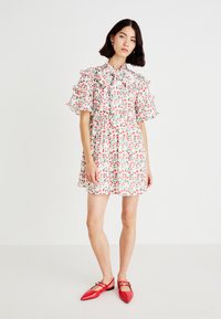 Vestido de manga corta con un cuello con volantes, adornado con un patrón floral de bayas rojas y hojas verdes sobre un fondo blanco. Llevado con zapatos rojos.