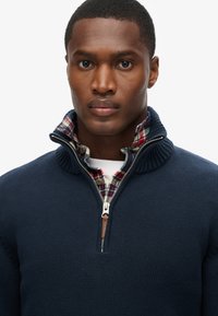 Marineblauer Strickpullover mit hohem Kragen und Reißverschluss, über einem karierten Hemd in Rot-, Grün- und Blautönen getragen. Strukturierte Stoffdetails sichtbar.