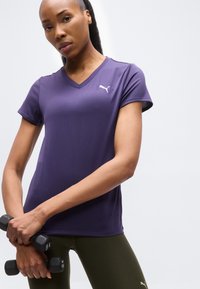 Γυναίκα με μωβ πουκάμισο Puma και ελιά κολάν, κρατώντας δύο μαύρα αλτήρες 1 κιλού μπροστά σε λευκό φόντο.