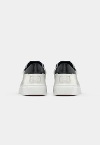 Adidași albi cu un tab de călcâi negru, exterior din piele netedă, talpă din cauciuc rezistentă și o etichetă de marcă pe spate.