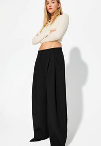 Schwarze, weit geschnittene, plissierte Hosen mit elastischem Bund, kombiniert mit einem beigen Langarmoberteil. Glatte Textur und minimalistisches Design.