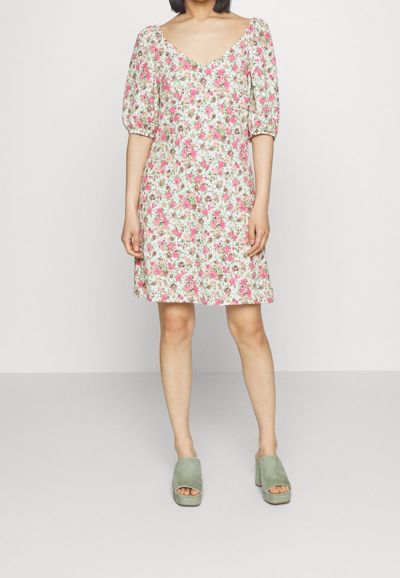 Femme portant une robe courte à manches bouffantes fleuries avec des fleurs roses et vertes, se tenant devant un fond uni gris clair.