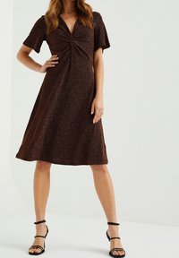 Brun, kortärmad klänning med v-ringning och vridningsdetalj fram. Tillverkad av textilerat tyg med en knälång, A-linjeformad silhuett.
