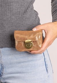 Metalen clutch in roségoud met een getextureerde afwerking, afgeronde vorm en sluiting met goudkleurig slot, vastgehouden in een hand tegen licht denim.