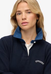 Pull-over bleu marine avec col en denim et patte de boutonnage blanche. Comprend une poche avant et le logo "SUPERDRY" brodé en blanc. Matière en coton.