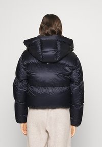 Marineblauwe pufferjack met een afneembare capuchon, voorzien van een gewatteerd design en elastische manchetten, gecombineerd met lichtgrijze broeken.