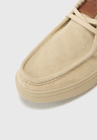 Chaussure en daim beige avec une finition texturée, présentant un bout arrondi et des lacets cirés. La semelle est plate et fabriquée dans un matériau de couleur claire.