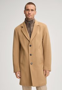 Cappotto beige in misto lana con rever a punta, design monopetto, tre bottoni e due tasche frontali; indossato sopra una giacca marrone trapuntata con colletto alto.