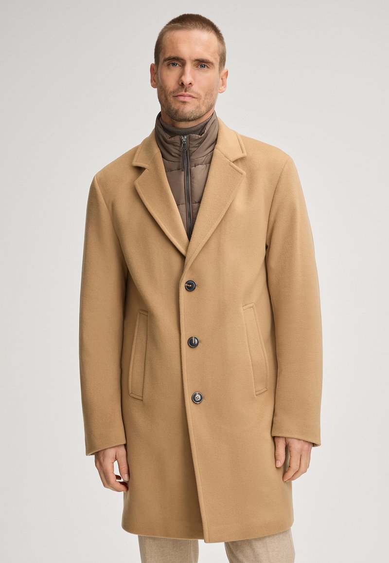 Cappotto beige in misto lana con rever a punta, design monopetto, tre bottoni e due tasche frontali; indossato sopra una giacca marrone trapuntata con colletto alto.