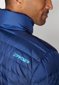 Doudoune bleu marine avec texture matelassée et col haut. Présente un logo "SPYDER" bleu contrastant sur l'épaule gauche.