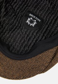 Crni kviltani materijal s smešastim rubom u uzorku riblje kosti, sa "Brixton" logom u beloj boji na crnoj zakrpi. Vidljive teksturirane pojedinosti.