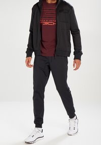 Schwarze Reißverschlussjacke mit Tasche, kastanienfarbenes T-Shirt mit Grafik, schwarze Jogginghosen und weiße Sportschuhe. Glatter Stoff, enganliegendes Design.
