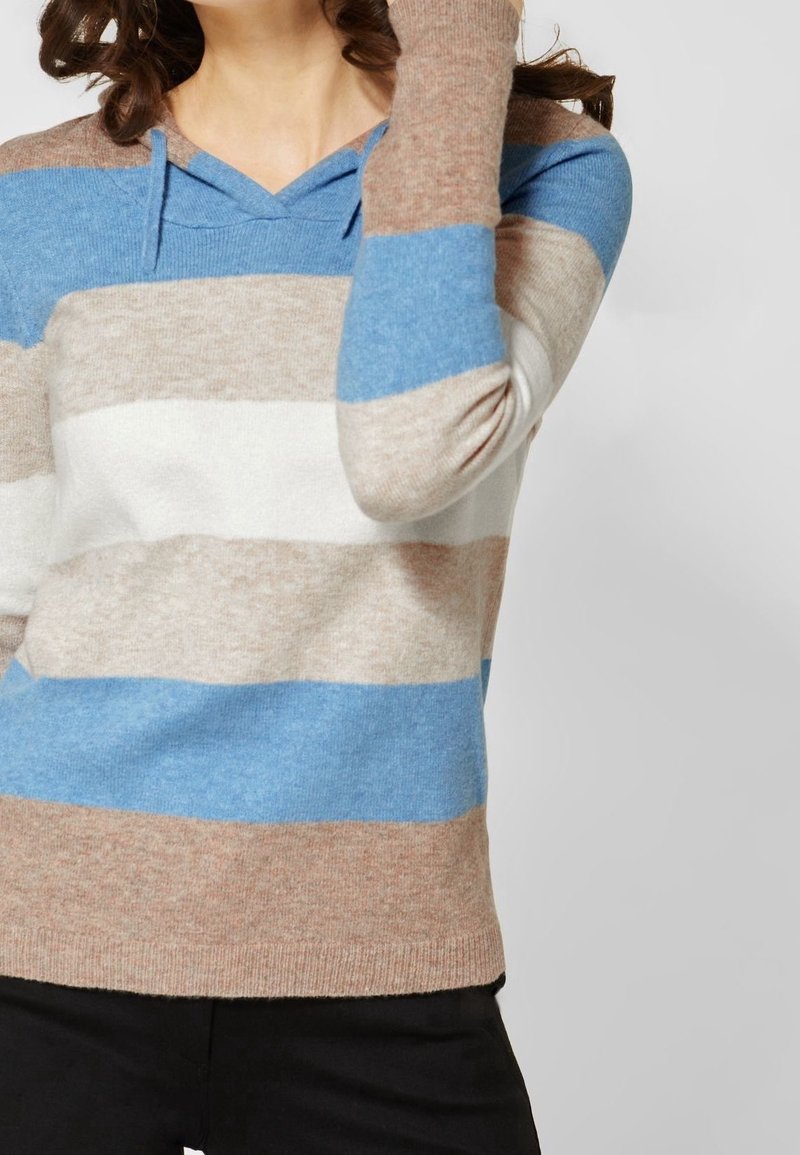 Femme portant un sweat à capuche à rayures horizontales beige, bleues et blanches, le bras levé près de ses cheveux, sur un fond uni.