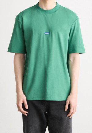 Grünes Baumwoll-T-Shirt mit Rundhals und kurzen Ärmeln. Auf der Vorderseite befindet sich ein kleines blaues "HUGO"-Logo. Lockere Passform, glatte Textur.