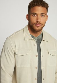 Camisa beige clara de botones con dos bolsillos en el pecho, llevada sobre una camiseta de cuello en V gris oscuro. Textura suave y diseño casual.