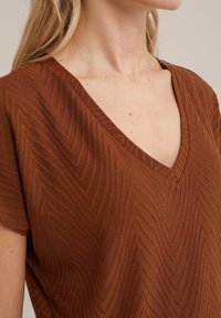 Brun v-ringad topp med ett strukturerat zigzagmönster, korta ärmar och en mjuk tygfinish. Detaljer inkluderar en något avslappnad passform.