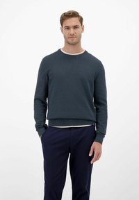 Dunkelgrüner Strickpullover mit Rundhalsausschnitt, gerippten Bündchen und Saum. Über einem helleren Hemd getragen und mit einer marineblauen Hose kombiniert.