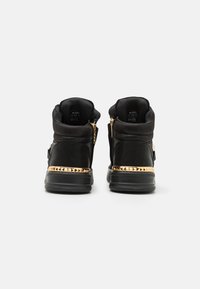 Svarta hög-top sneakers med en texturerad tyg, guldspikade detaljer runt sulan och en dragkedja på sidan.