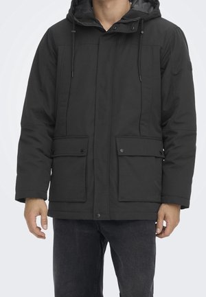 Uomo che indossa una giacca invernale nera con cappuccio, tasche frontali con pattina e coulisses regolabili, abbinata a jeans scuri.