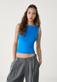 Giovane donna con lunghi capelli castani che indossa un top senza maniche di un brillante blu e pantaloni grigi larghi e plissettati, in piedi con le mani nelle tasche.