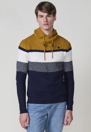 Un joven está de pie vistiendo un suéter de punto con bloques de color y cuello alto, que presenta franjas en mostaza, blanco, gris y azul marino, y jeans de un tono azul claro.