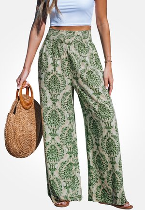 Femme portant un pantalon taille haute à jambes larges avec des motifs floraux verts, un crop top blanc, des sandales marron clair, et tenant un sac à main rond en osier.