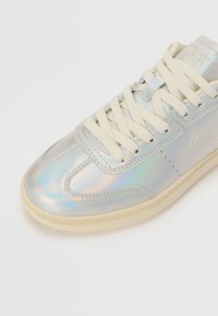 Sneaker métallique avec un dessus lisse et irisé, des lacets crème et une semelle en caoutchouc de couleur claire. Présente des détails de marque subtils sur le côté.
