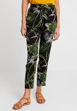 Kvinde iført sorte slim-fit bukser med grønne og lilla tropiske bladprint, kombineret med tanfarvede sandaler og en gul top.