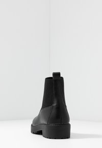 Botín negro de cuero con parte superior de elastano estriado y tira de agarre, colocado sobre una superficie blanca con un fondo claro liso.