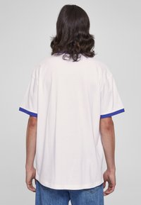 Hvit unisex t-skjorte med korte ermer, med blå detaljer rundt mansjettene, i en avslappet passform. Glatt bomullstekstur, enkelt design.