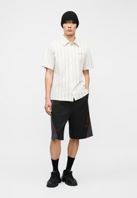 Νέος άνδρας που φοράει μαύρο σκουφάκι, λευκό κοντομάνικο ριγέ πουκάμισο, μαύρο σορτς με πλαϊνές λεπτομέρειες, μαύρες κάλτσες και μαύρα αθλητικά παπούτσια, στέκεται όρθιος.