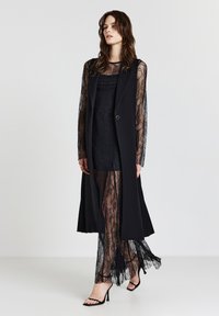 Langer schwarzer Weste mit durchsichtigen, langen Spitzenärmeln, layered über einem figurschmeichelnden Spitzenkleid mit zarten Mustern und einer fließenden Silhouette.