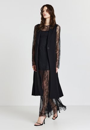 Gilet lungo nero con maniche lunghe in pizzo trasparente, sovrapposto a un abito aderente in pizzo, caratterizzato da motivi delicati e una silhouette fluida.