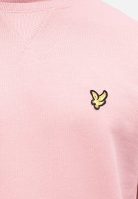 Sweatshirt rosa com acabamento texturizado, apresentando um pequeno logótipo de águia bordado a preto e dourado no peito. Gola canelada.