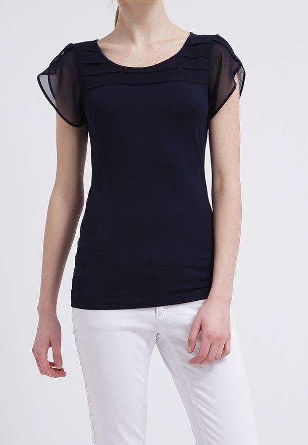 Mörkblå topp med genomskinliga, korta puffärmar och lagerdetaljer vid halslinningen. Bärs med vita byxor, slät tygtextur.