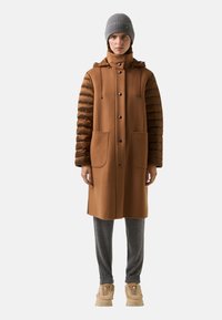 Hnědý kabát z vlněné směsi s polstrovanými hnědými rukávy, zapínání na knoflíky, dvěma předními kapsami a vysokým límcem, v kombinaci se šedými kalhotami a pletenou čepicí.